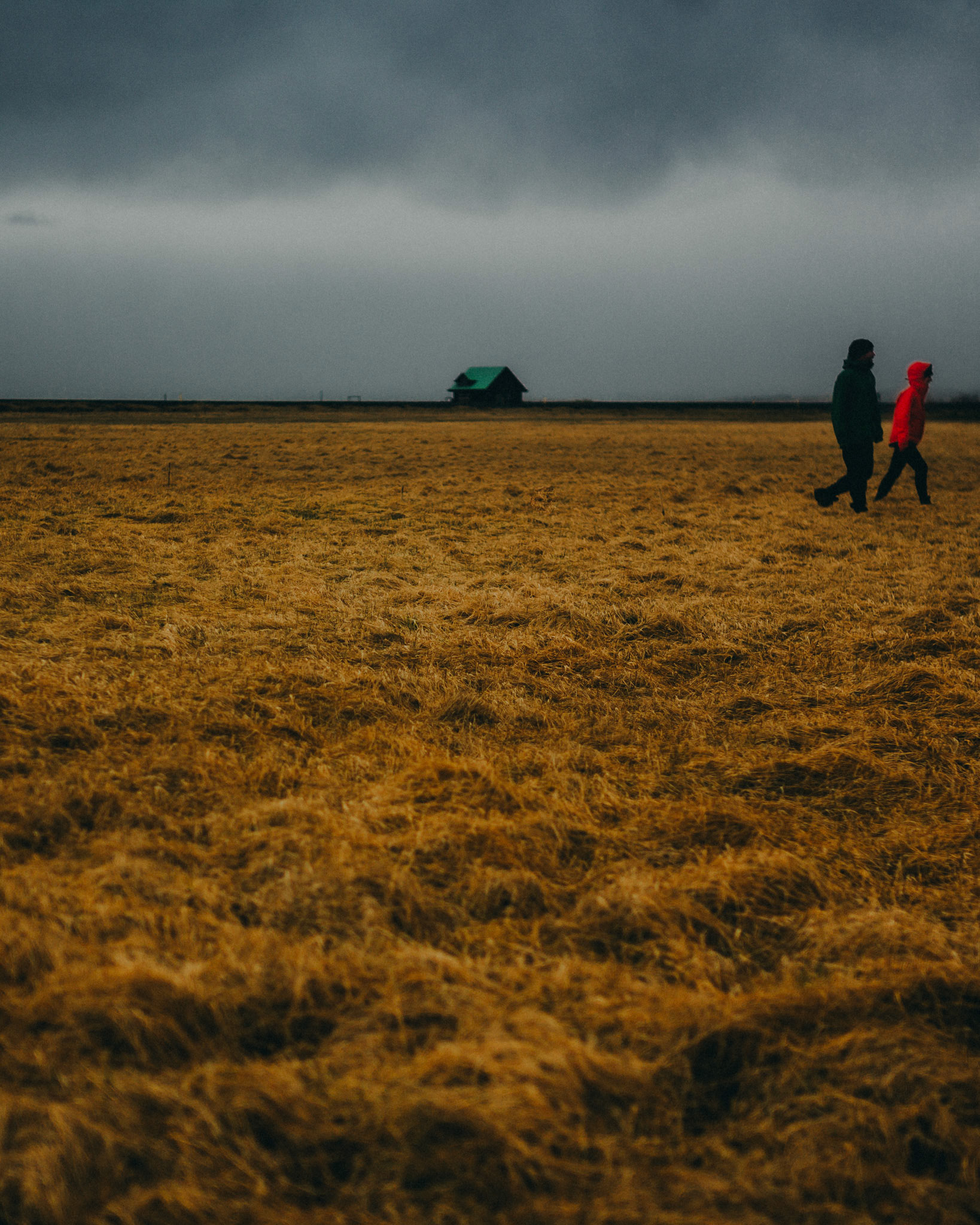 A minimalist landscape photo of a lone cabin and two people walking in an open field in Skógar, Iceland, May 2016, Leica M.