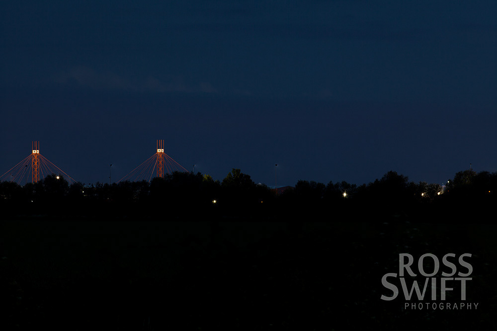 A dark suburban landscape at twilight, with two illuminated red structures and street lights glistening through the trees.
