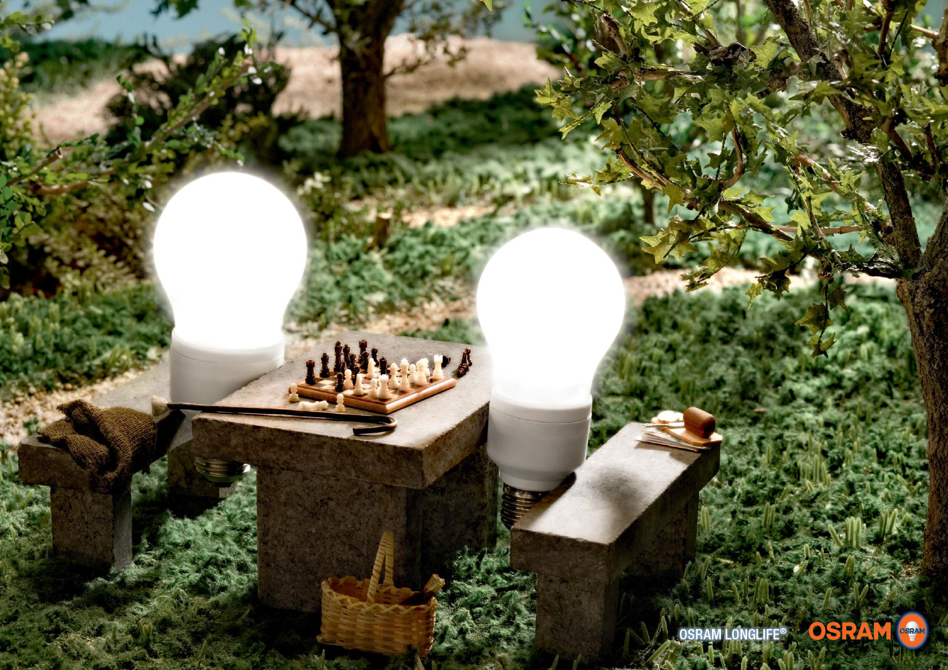 Langlebige Gluehbirnen sind umweltfreundlich. Zwei Gluehbirnen von Osram sitzen mit Deckchen und Picknick-Korb im Park und spielen Schach.