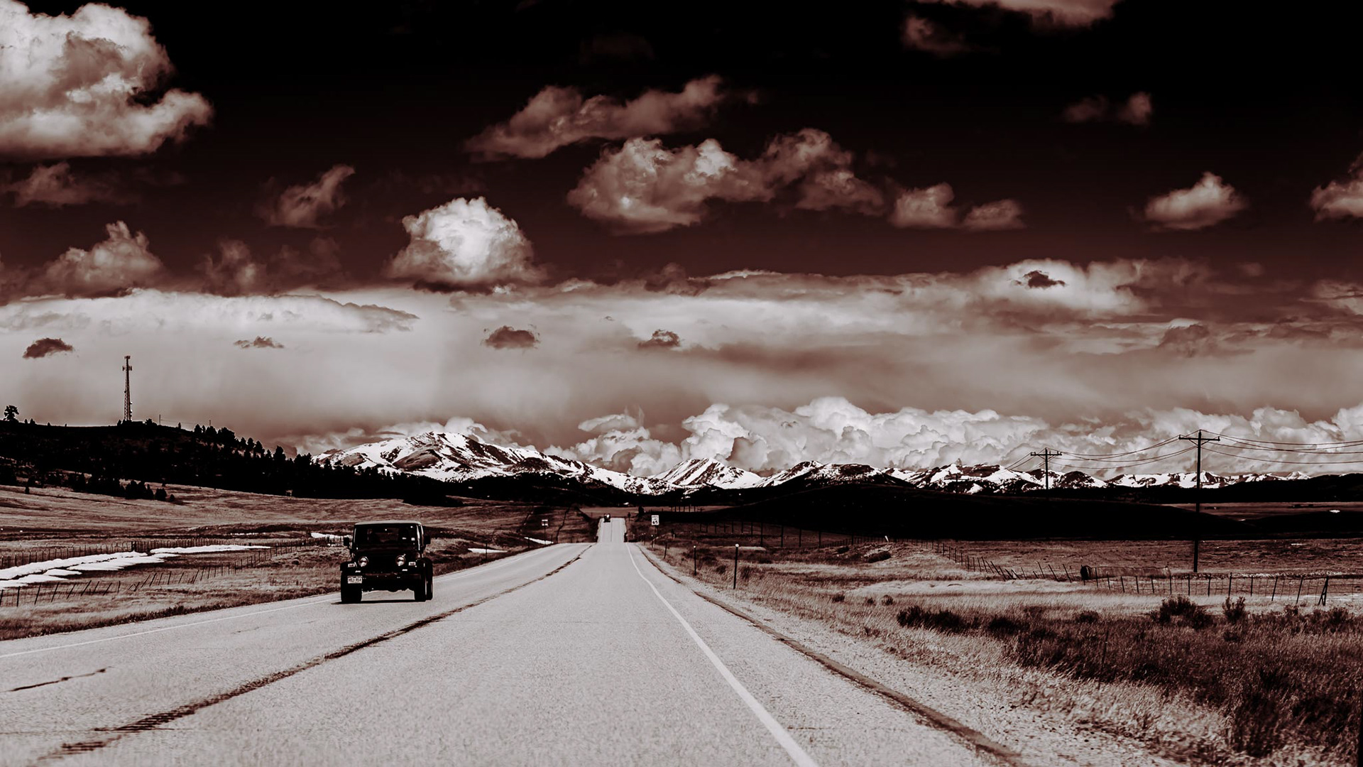 Vintage Black and White, Top to Bottom, darker sky, then white clouds, filing down behind snowy rocky mountains of colorado, on an open road and a jeep passing on road.