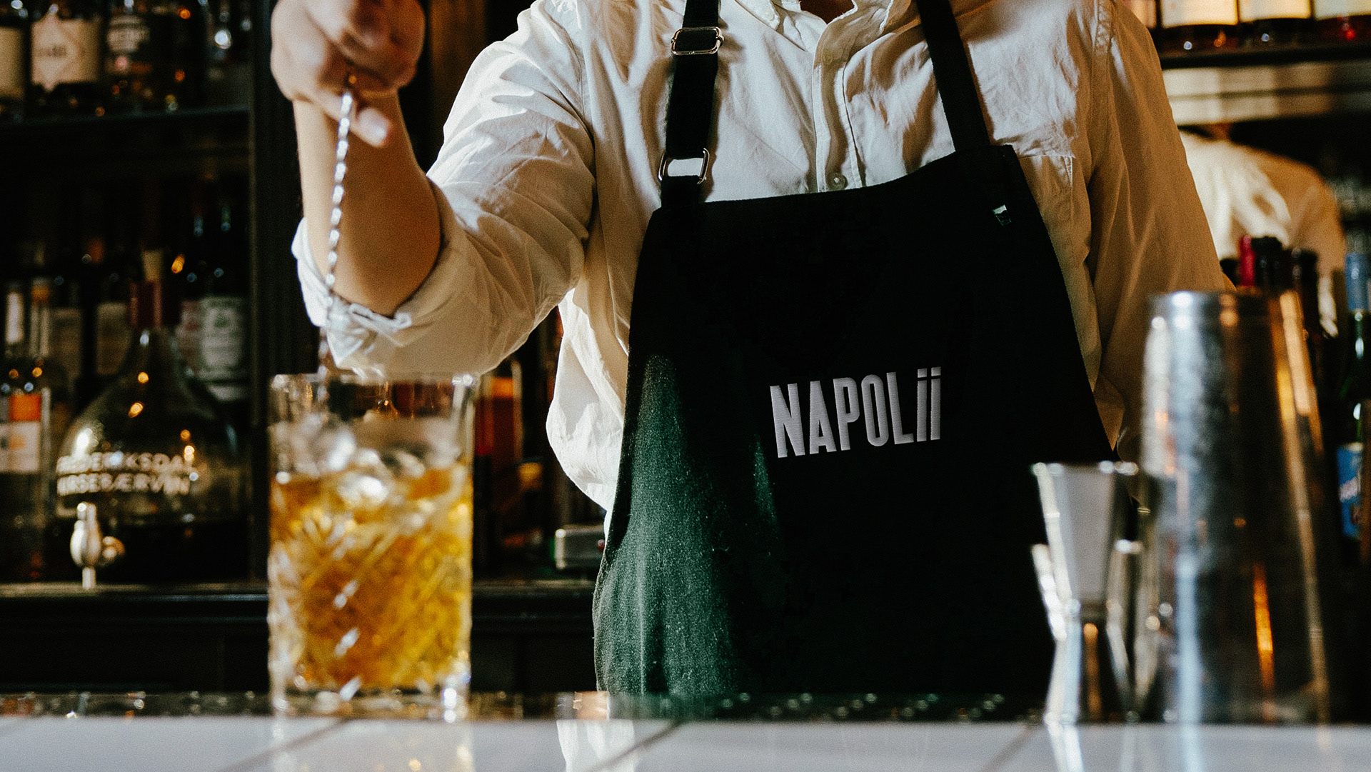 Man wearing a dark green apron with embroidered Napolii logo preparing a cocktail