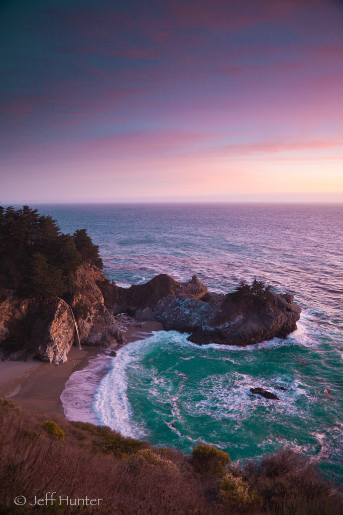 A waterfall falling onto a beach in a cove at sunset