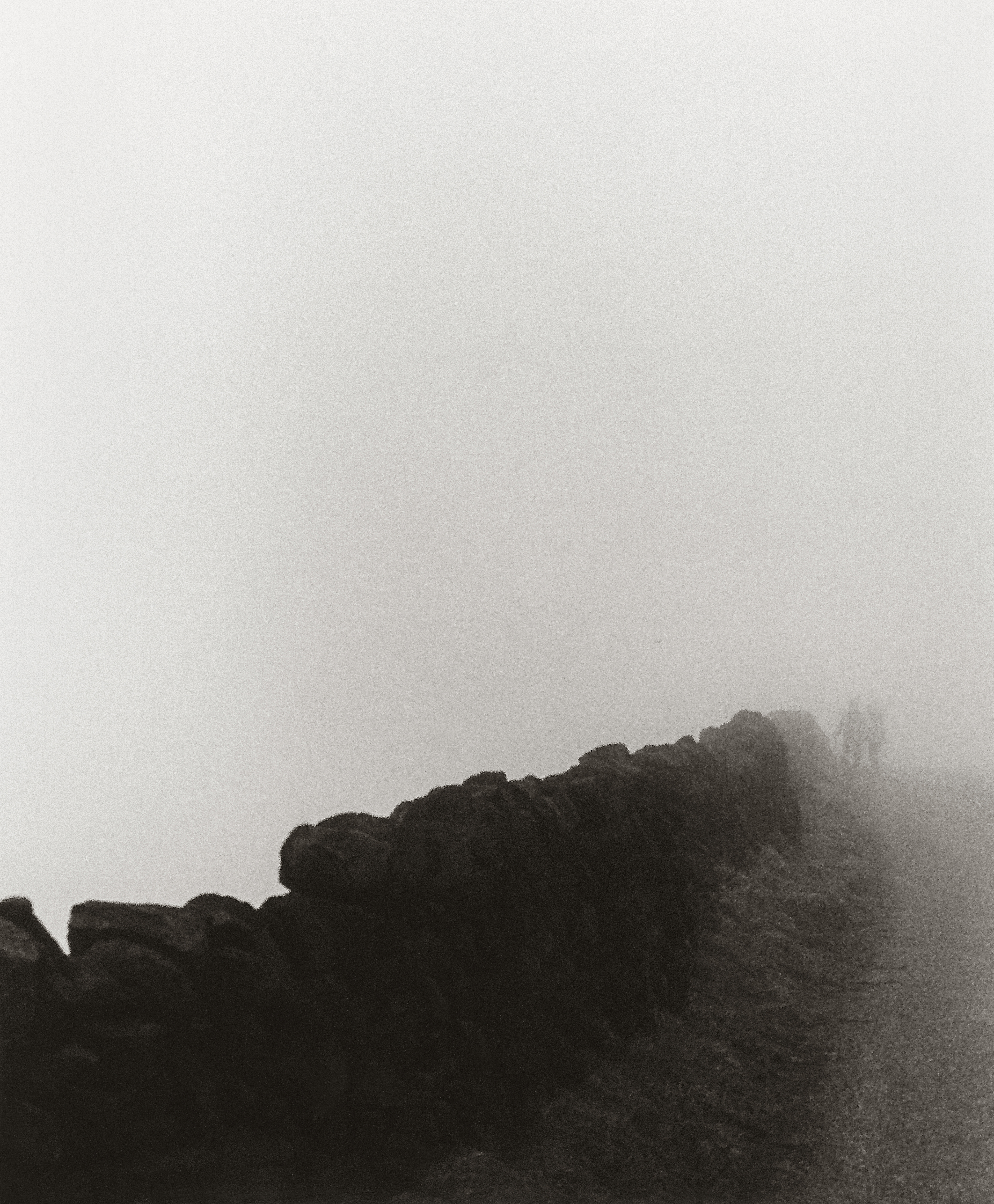 A very foggy country road/path with a small stone wall along it with 2 people just noticeable in the distance