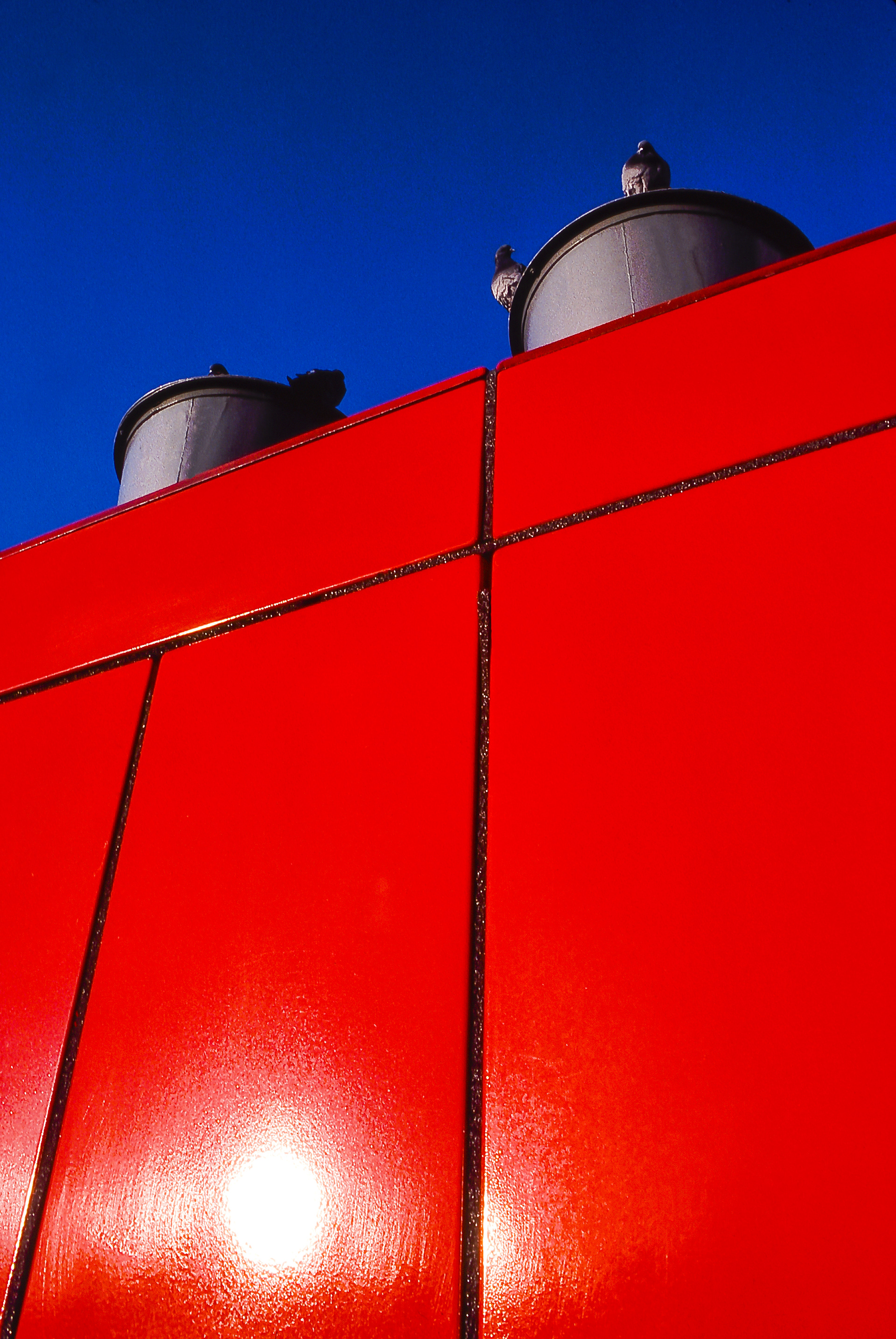 Vivid red metal wall with pigeons on  grey round chimneys against a blue sky