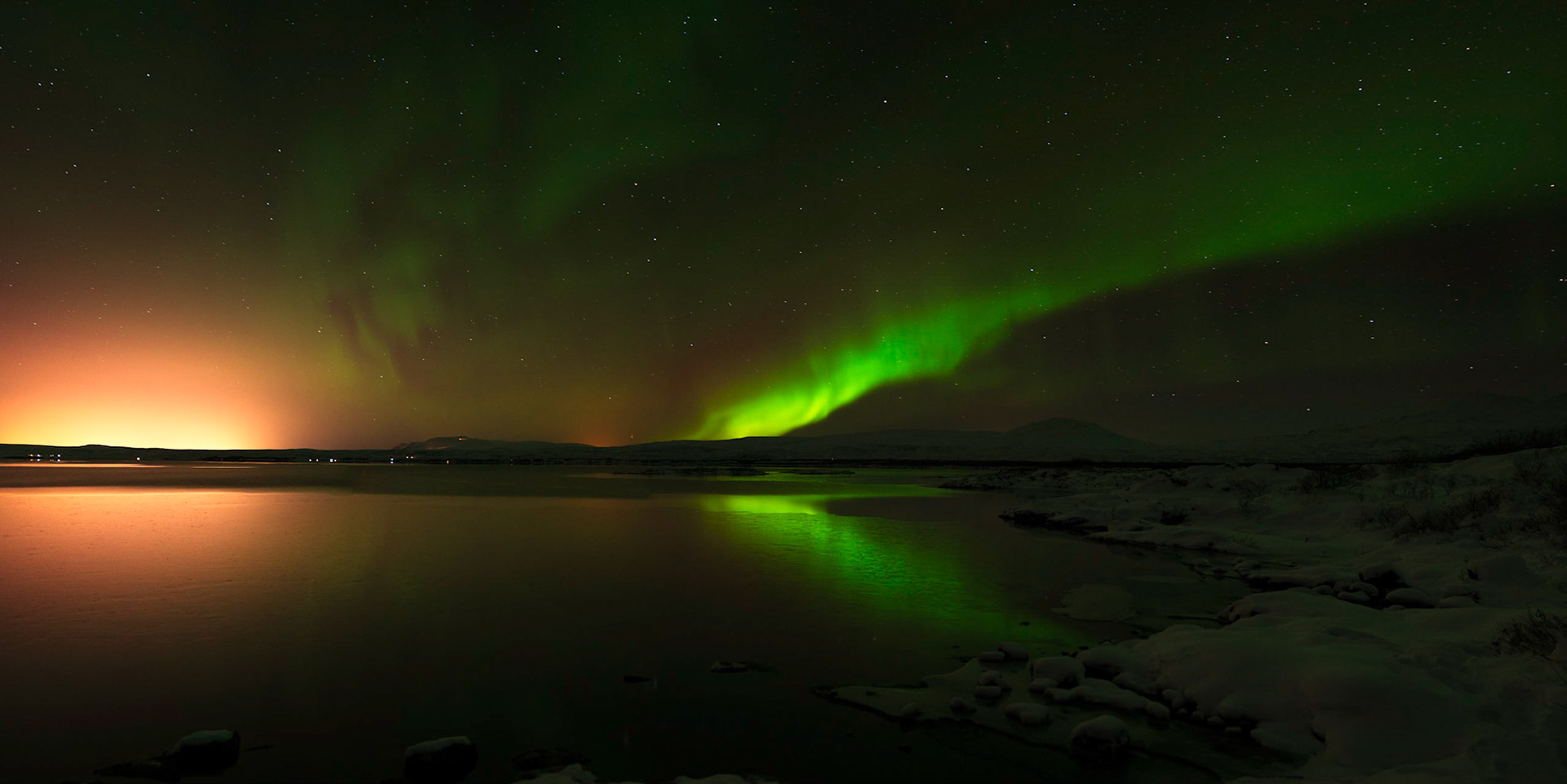 The northern lights over Þingvallavatn, in Þingvellir National Park, Iceland.  The orange glow on the left is coming from Reykjavik.Þingvellir National ParkJanuary 31, 2016This is a panoramic images consisting of 11 frames stitched in Photoshop. Additional processing in Lightroom and Photoshop.PENTAX K-3, Sigma 18-35mm f/1.8 DC HSM ArtISO 400 18 mm  20.0 sec at ƒ / 1.8