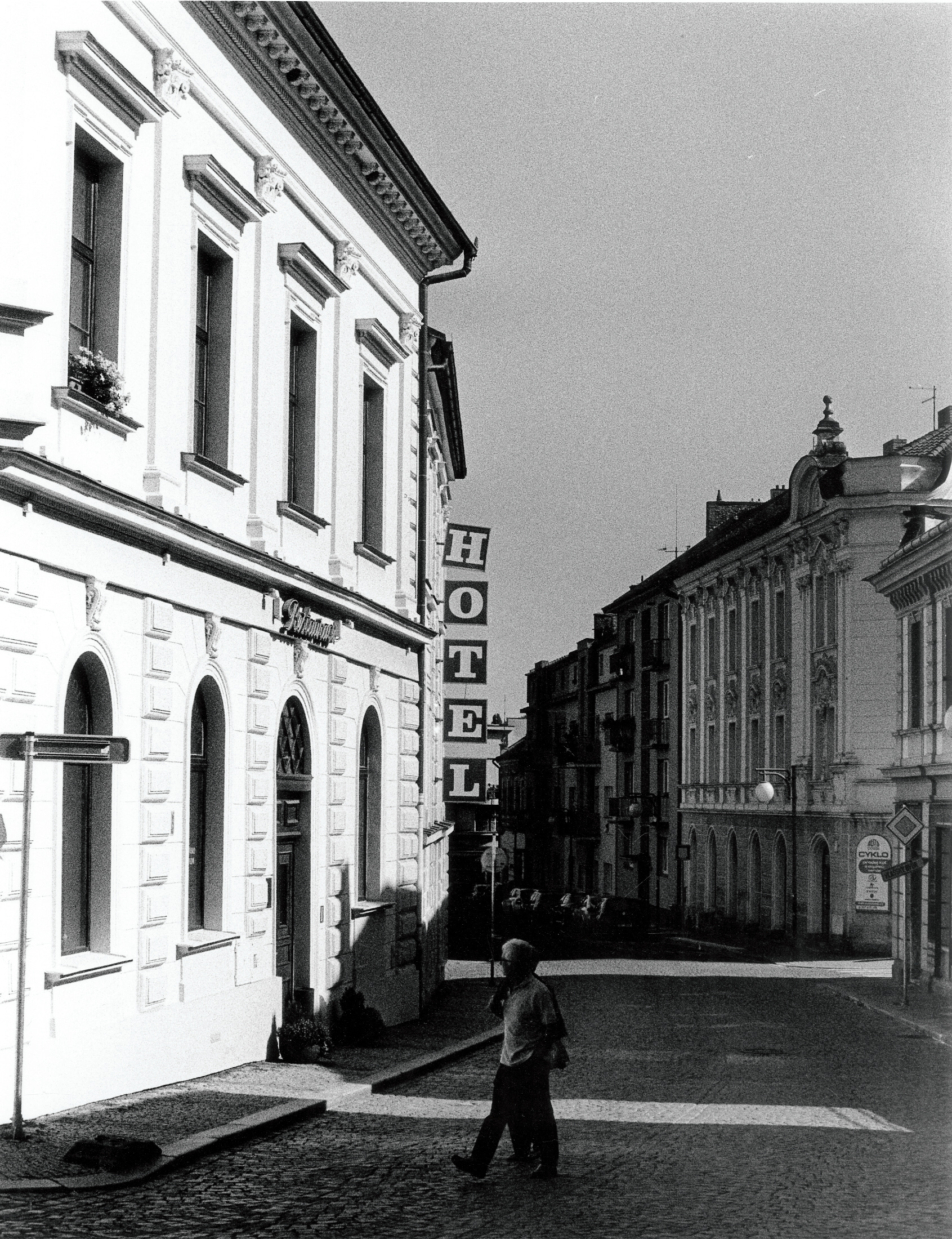 a couple cross the street in Kútna Hora. Behind them the vertical sign for a local hotel has a crooked 'H'