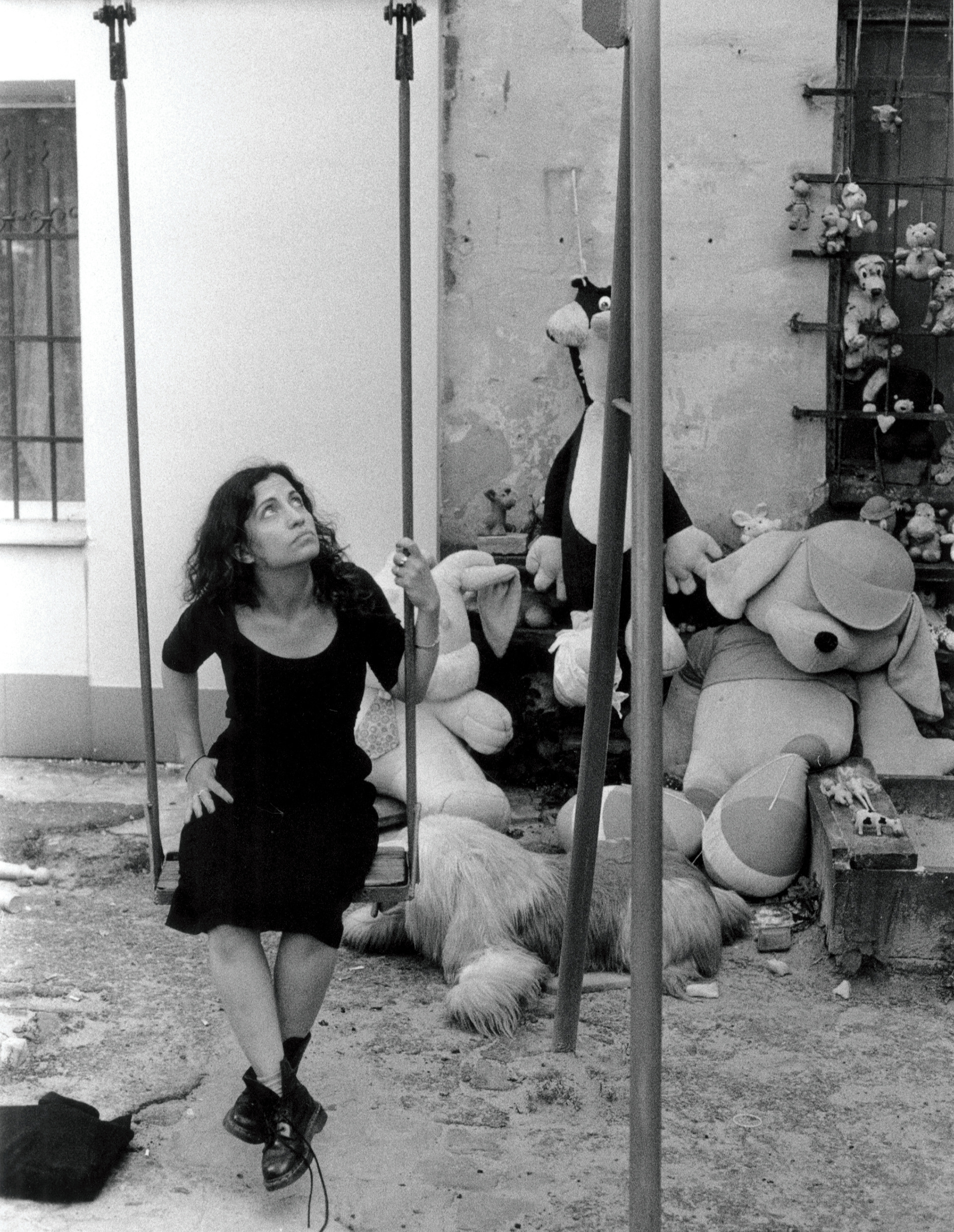 Ana looks up while perched on a swing, in the background is an assortment of large stuffed toys