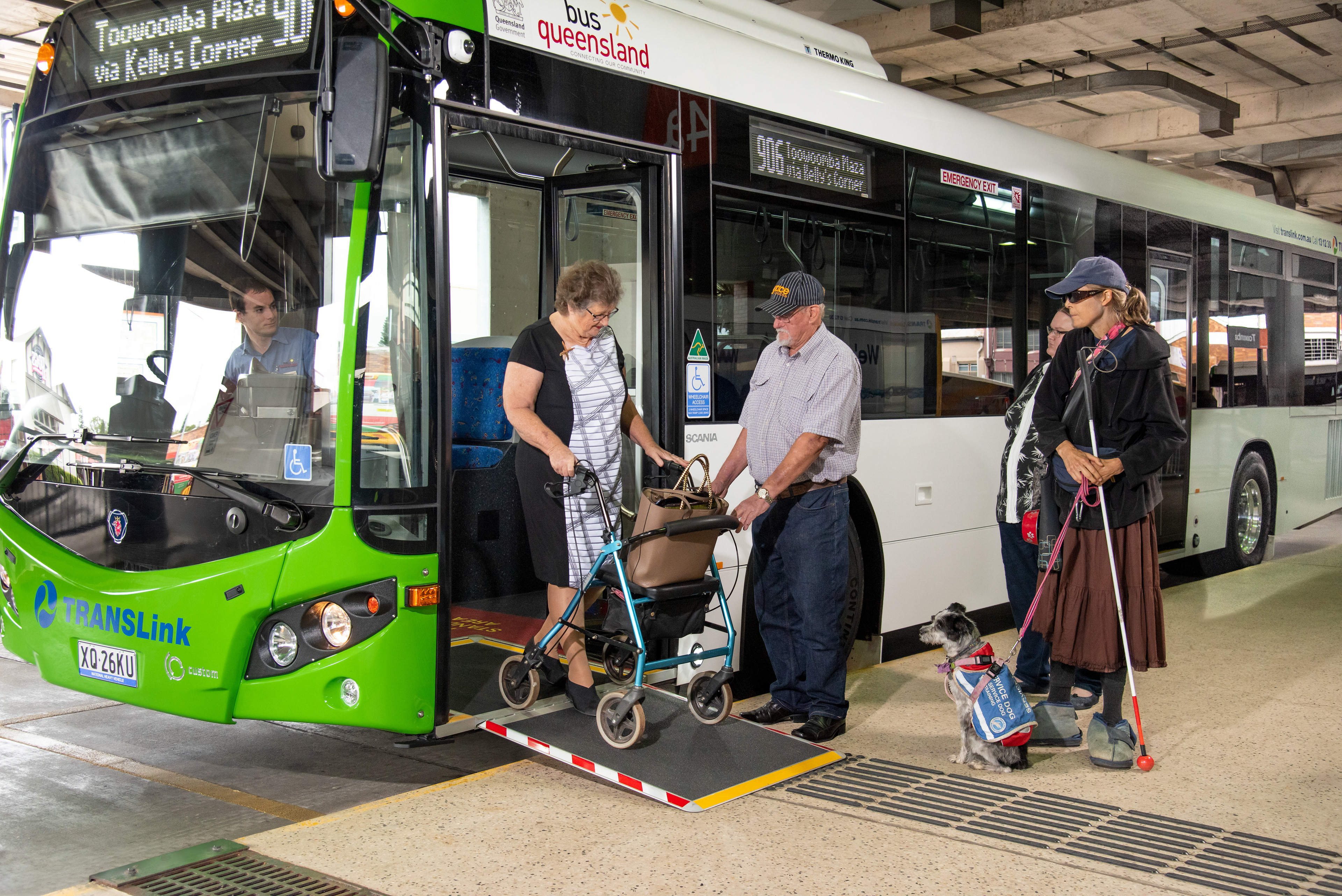 Toowoomba bus passengers with walker being assisted off the bus while a blind lady with her dog watching on.