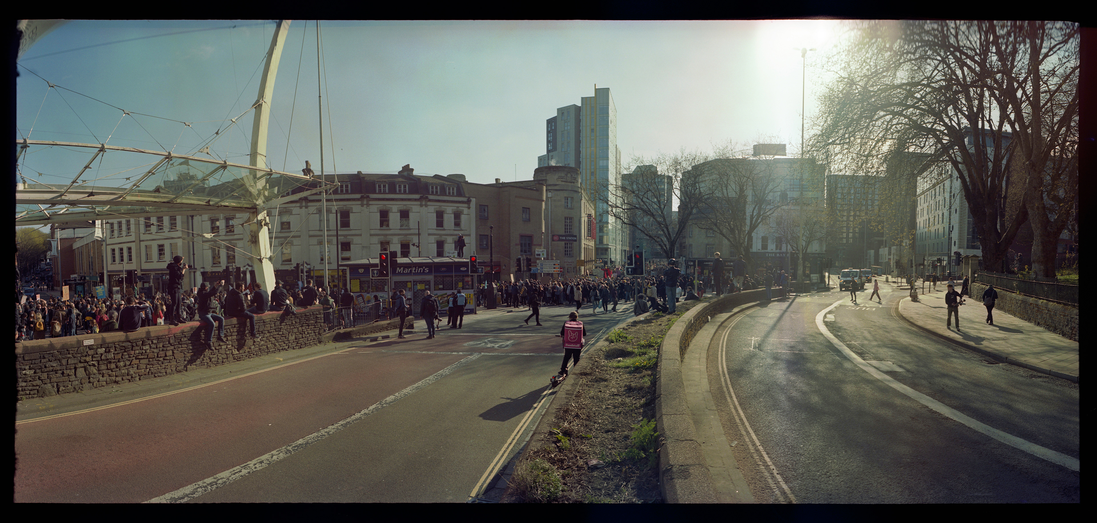 The moment I caught up with the protest march, a wide angle view of Bristol city centrey, with many protesters marching in the streets from right to left. The sun is above and behind them.