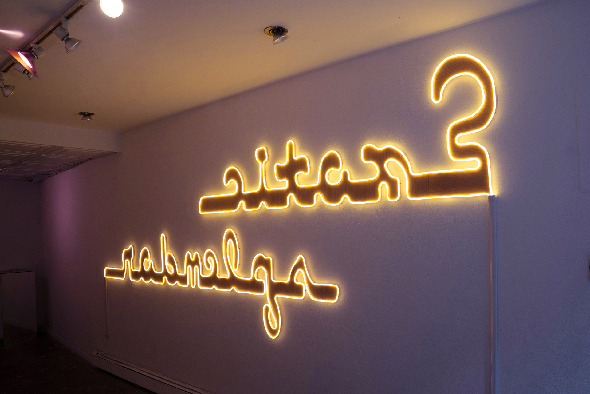 A yellow neon sign with script text on a pink wall