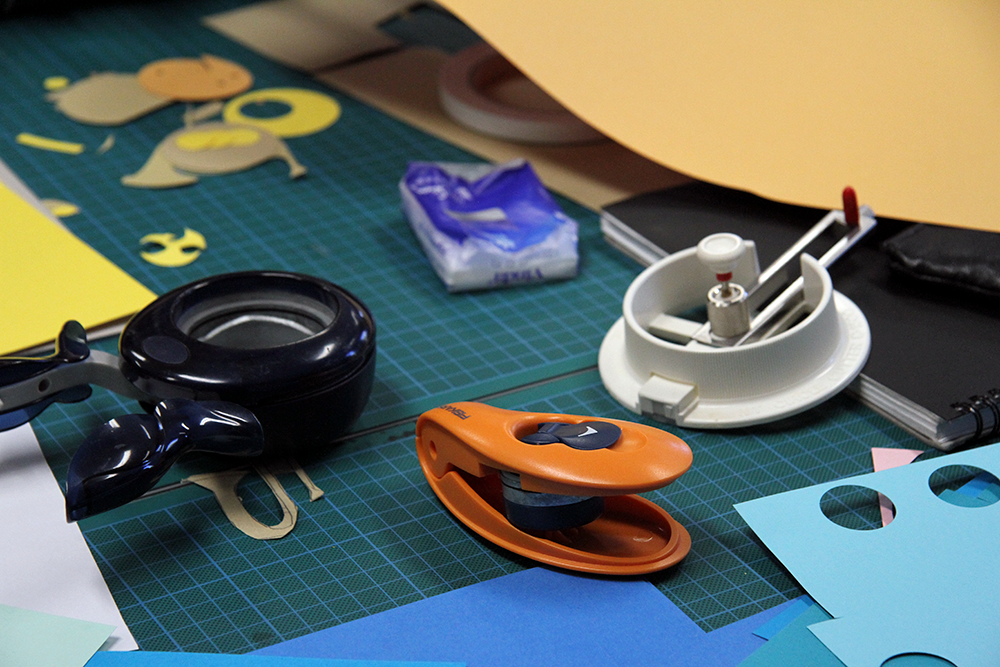cutting mats on a desk, covered in circles of brightly coloured paper, that have been cut out using oversized hole-punches and circle-cutting tools..