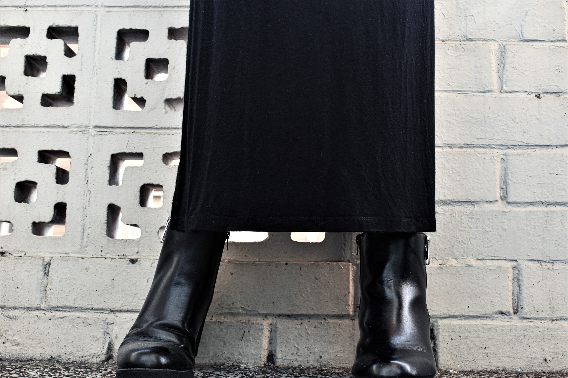 Kerry is wearing black boots with a long black skirt, poed against a white brick wall.