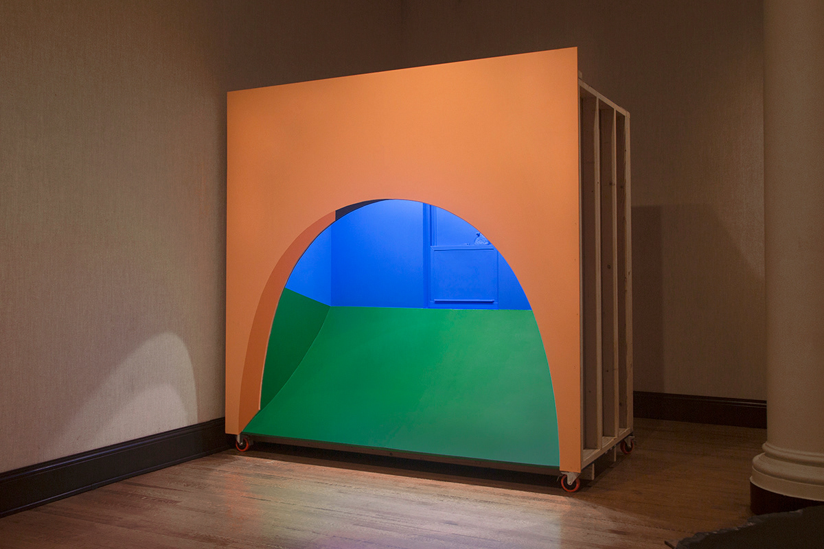 Angled view of the sculpture "Paradise of Greenery." An 8 foot high by 8 foot long by 4 foot deep room on wheels. viewed from the front, there is a green ramp that reaches from the front bottom edge of the room to the middle of the back wall. The top section of the room has a window with a ceramic cactus on the ledge. The entire top half of the room is painted blue. With the green ramp and the blue wall, the room looks like a landscape of green pastures and blue skies. The entrance to the room is a large sheet of plywood in the shape of an orange archway. The sculpture was created by imagining what a mural on the Israeli Separation Barrier would look like in 3 dimensions