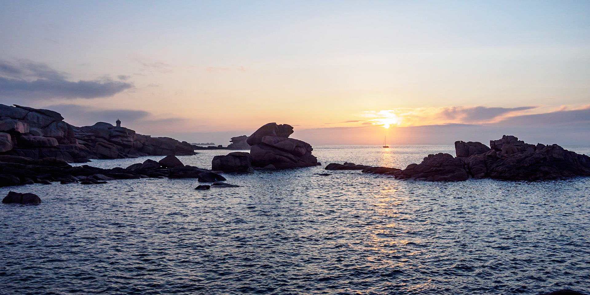 The slightly veiled sun setting over the English Channel at Ploumanach, France, reflecting in the water,  casting a soft dim light over the pink granite rocks. And right under the light of the sun, a small baot passed us by.
