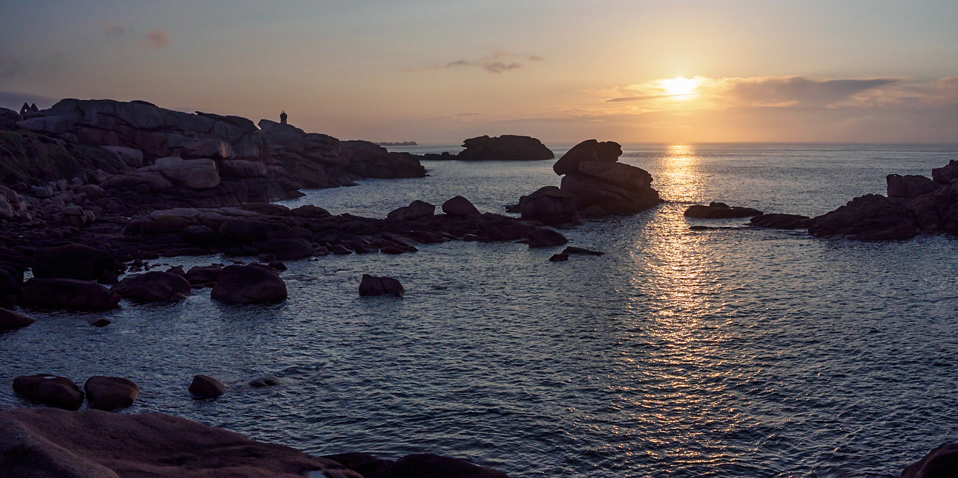 The slightly veiled sun setting over the English Channel at Ploumanach, France, reflecting in the water,  casting a soft dim light over the pink granite rocks, the light house of Mean ruz and the remains of an old house.